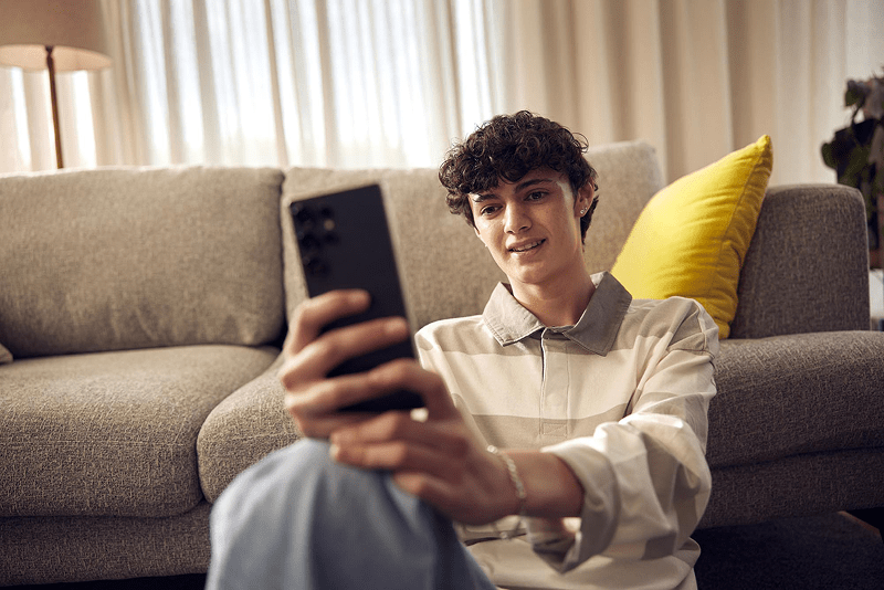 Boy sitting against a grey couch holding his phone out in front of him and looking at it