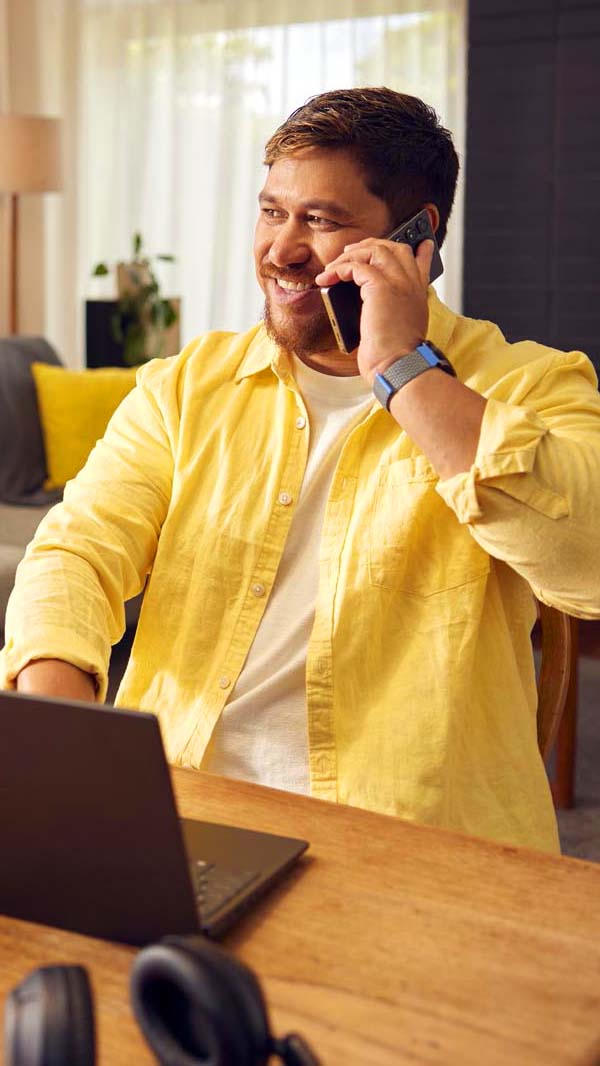 Man wearing yellow shirt sits at a table talking on his mobile phone with a laptop open in front of him.