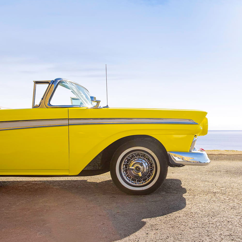 A yellow Ford Fairlane parked in front of a beach.