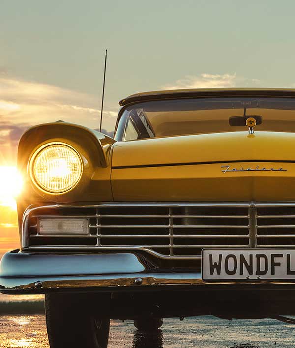 A yellow Ford Fairlane with headlights on and a number plate that says 'WONDFL' parked on a beach as the sun sets.